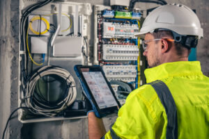Technicien électricien consultant une tablette devant un tableau de fusibles dans un atelier industriel.